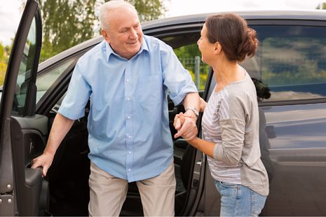 A caregiver assists an older man as he steps out of a car, suggesting transport or mobility support &mdash; reflecting independence and everyday assistance.
